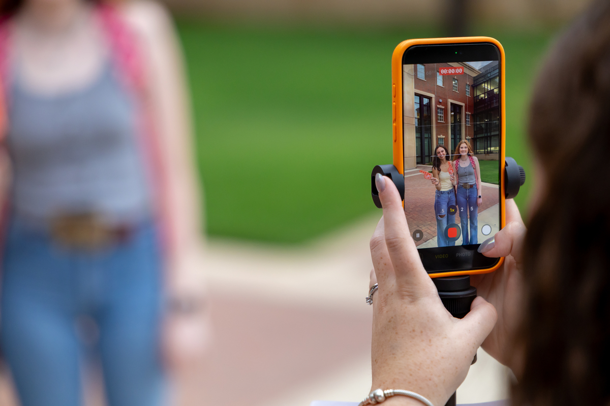 A person records two students standing together outside a brick building with a smartphone.