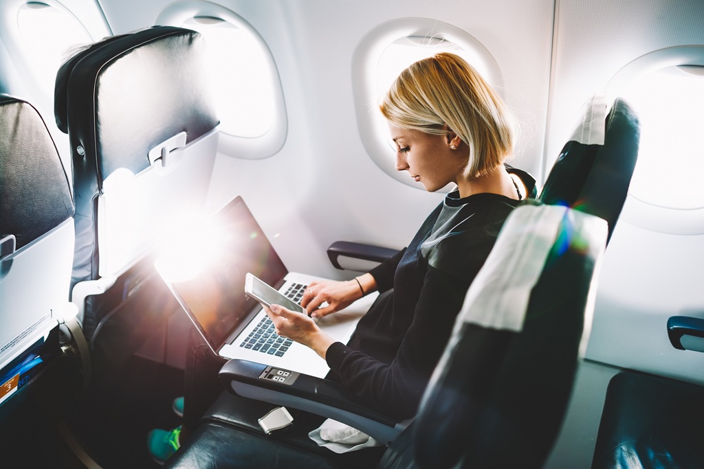 A person sits on an airplane using a laptop and phone while sunlight shines through the window beside them.