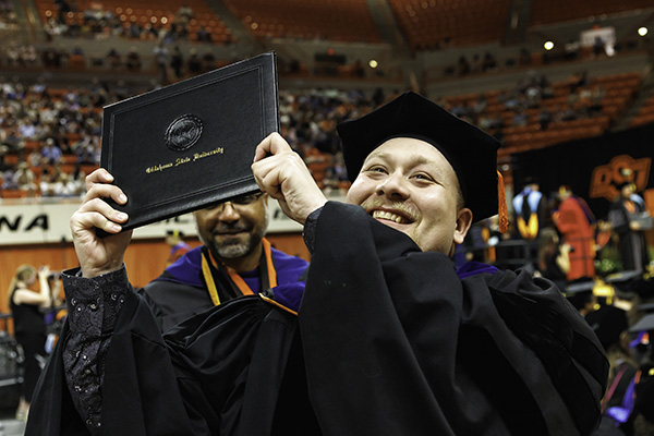 A man smiles while holding his diploma at graduation.